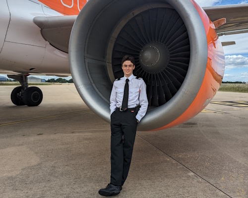 Tom standing in front of an Airbus A320 engine on the airport ramp; aircraft fuselage and tarmac visible in daylight; subject appears proud and confident; no visible text