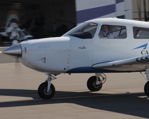 Tom seated at the controls of a small single-engine airplane during his first solo flight wearing a headset and hands on the yoke; daytime runway and open sky visible through the windshield; cockpit instruments visible; mood proud and focused; no visible text