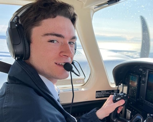 Tom seated in the left cockpit seat of a Piper Seneca V, wearing a headset with hands on the yoke while flying during flight training; instrument panel and sky visible through the windshield, cockpit interior of a twin-engine light aircraft; mood focused and determined; no visible text